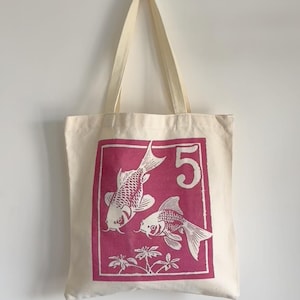May include: Cream-colored tote bag with a magenta rectangular graphic featuring two koi fish and the number 5. The bag is 15 inches (38cm) high, 14 inches (35cm) wide, and has a 23-inch (60cm) handle. The bag is hanging against a white wall.
