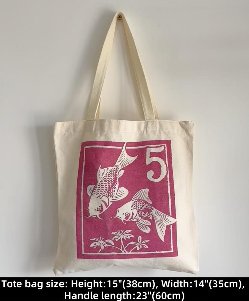 May include: Cream-colored tote bag with a magenta rectangular graphic featuring two koi fish and the number 5. The bag is 15 inches (38cm) high, 14 inches (35cm) wide, and has a 23-inch (60cm) handle. The bag is hanging against a white wall.