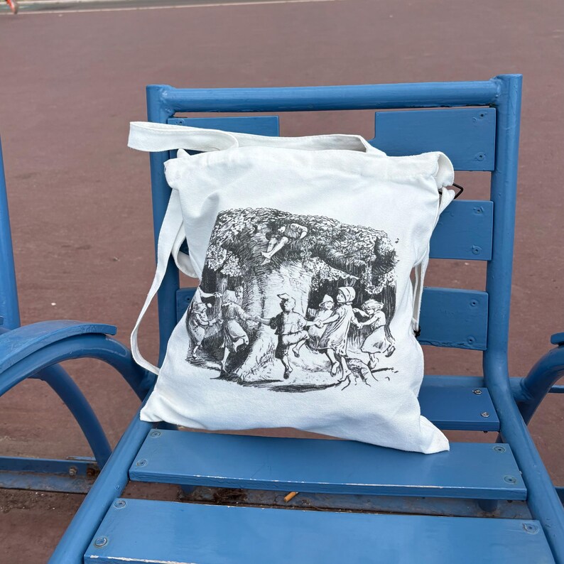 May include: A white tote bag with a black and white illustration of people dancing in a forest. The bag is sitting on a blue bench. The bag has long white straps.