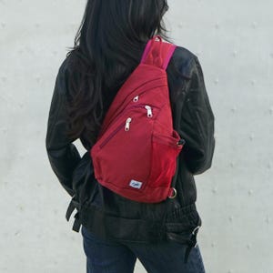 May include: A red sling backpack with multiple zippered compartments and a mesh side pocket. The bag is worn over the shoulder and across the chest. The bag has a small white tag with a blue design.