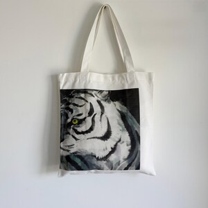 May include: White tote bag featuring a watercolor painting of a tiger's face. The tiger has black stripes, a white face, and bright yellow-green eyes. The bag has two white handles and is hanging against a white wall.