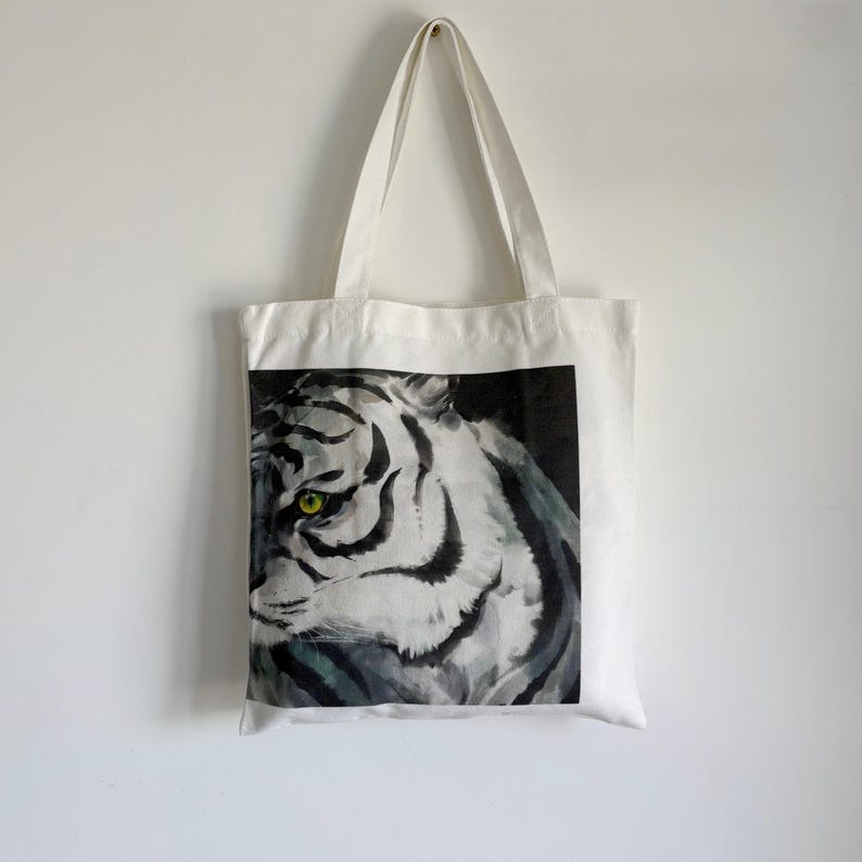 May include: White tote bag featuring a watercolor painting of a tiger's face. The tiger has black stripes, a white face, and bright yellow-green eyes. The bag has two white handles and is hanging against a white wall.