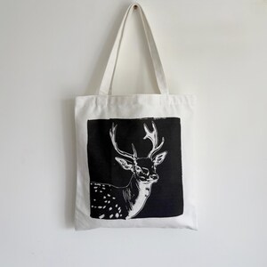 May include: White canvas tote bag featuring a black and white illustration of a deer with large antlers. The deer is set against a black rectangular background. The bag has two white handles and is hanging against a white wall.