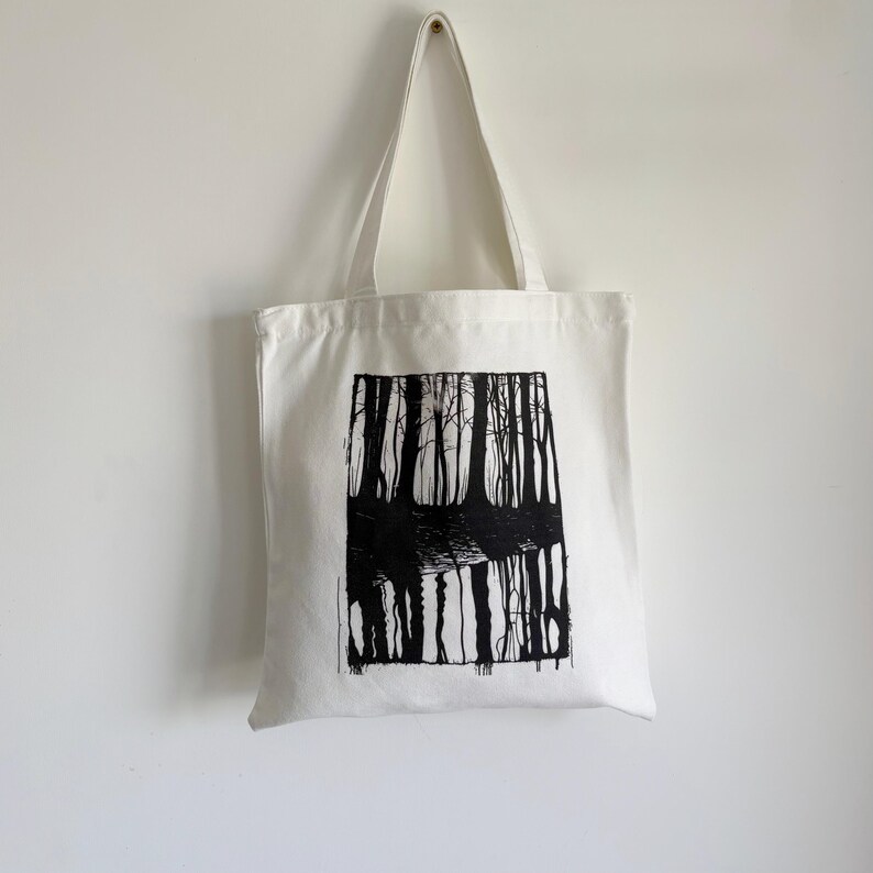 May include: A white canvas tote bag featuring a black and white forest scene. The artwork depicts tall trees reflected in water. The bag has long handles for carrying over the shoulder. The tote bag is hanging against a white wall.