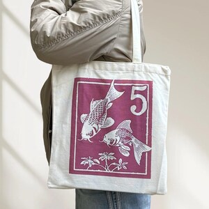 May include: A white tote bag featuring a magenta rectangular design with two koi fish and the number 5. The fish are white with detailed scales and fins. The bag is held by a person wearing a tan jacket.
