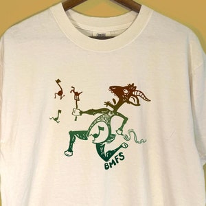 May include: White t-shirt with a graphic of a goat-like creature playing a drum. The illustration is in shades of green and brown, with musical notes and the letters "BMFS" visible. The shirt is hanging on a wooden hanger.