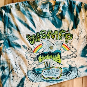 May include: A tie-dye t-shirt with a teal and cream pattern. The shirt features a graphic with the text "WSMFP" and a colorful design of a rainbow, mountains, and waves. The text "Asheville, NC July 24, 25, 26, 2025" is also visible.