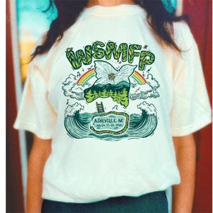 May include: Cream-colored t-shirt featuring a colorful graphic design. The design includes the text "WSMFP" in green, a rainbow, clouds, and a stylized map of a mountain range. The text "Asheville, NC July 24, 25, 26, 2025" is also included.