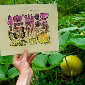 May include: A linocut print of a garden harvest with a yellow pumpkin, carrots, corn, a watering can, a butterfly, and other garden vegetables and flowers. The print is on a cream-colored paper and has the text "Garden Harvest!" at the bottom.