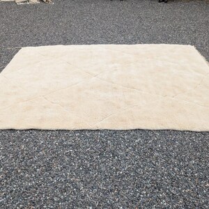 May include: A rectangular, cream-colored rug with a diamond pattern. The rug is made of a soft, plush material and is placed on a dark, textured surface. The rug's design features subtle, raised lines.