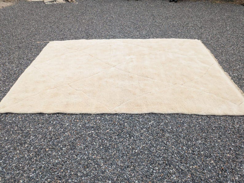 May include: A rectangular, cream-colored rug with a diamond pattern. The rug is made of a soft, plush material and is placed on a dark, textured surface. The rug's design features subtle, raised lines.