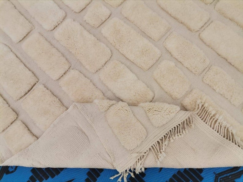 May include: A cream-colored rug with a brick pattern. The rug has a fringe border and is made of a soft, fluffy material.