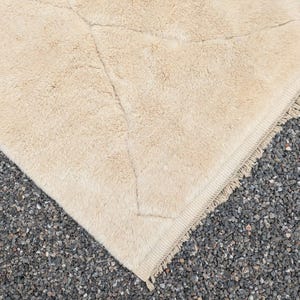 May include: A corner of a beige, textured rug with a geometric pattern. The rug has a soft, plush appearance and a fringed edge. The rug is placed on a surface of small, dark gray pebbles.