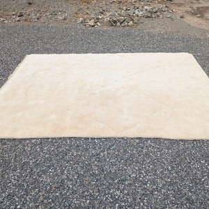 May include: A rectangular, beige rug with a diamond pattern. The rug is made of a soft, plush material and is placed on a gravel surface. The rug is a neutral color and would complement various interior design styles.