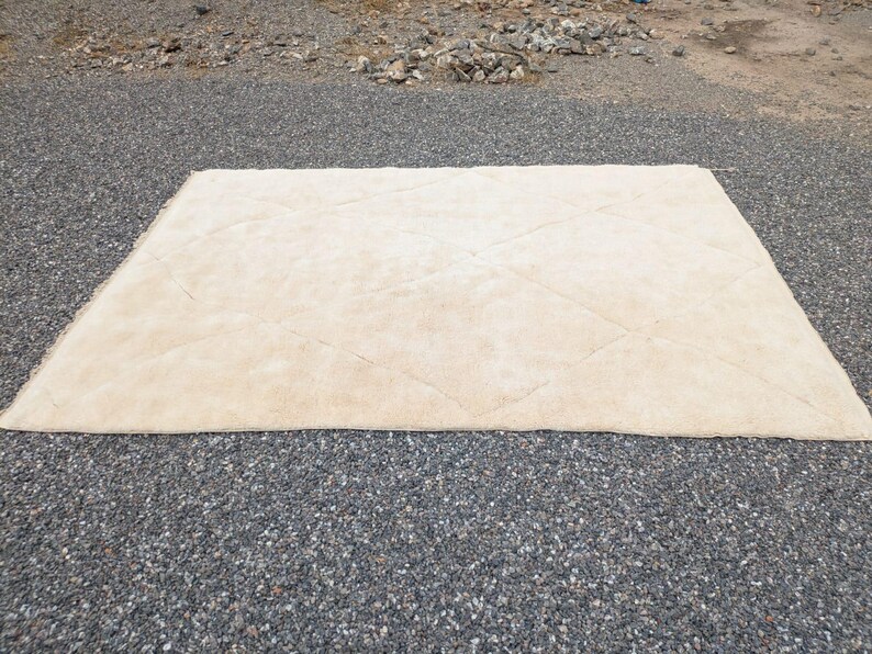 May include: A rectangular, beige rug with a diamond pattern. The rug is made of a soft, plush material and is placed on a gravel surface. The rug is a neutral color and would complement various interior design styles.