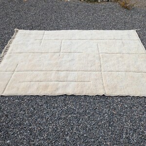 May include: A rectangular, off-white rug with a geometric design. The rug features a series of raised, rectangular and linear patterns across its surface. The edges have a fringed detail. The rug is placed on a bed of small, dark gray pebbles.