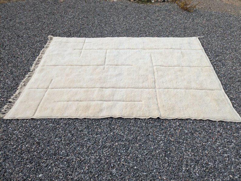 May include: A rectangular, off-white rug with a geometric design. The rug features a series of raised, rectangular and linear patterns across its surface. The edges have a fringed detail. The rug is placed on a bed of small, dark gray pebbles.