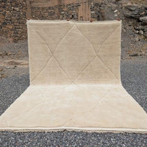 May include: A large, rectangular beige rug with a diamond pattern. The rug is displayed outdoors on a gravel surface, with a stone wall in the background. The rug's design features intersecting lines creating diamond shapes.