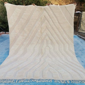 May include: A white handwoven rug with a geometric pattern. The rug has a fringe border and is being held up by two hands.