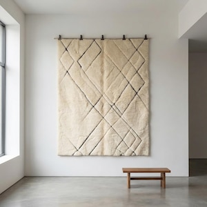May include: A large, cream-colored, geometric wall hanging with black lines, mounted on a wall. A small, wooden bench sits below it. The rug has a diamond pattern.