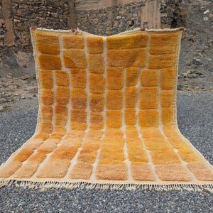 May include: A rectangular, hand-woven rug with a repeating pattern of golden-yellow and orange rectangular blocks. The rug has a cream-colored fringe along the bottom edge. The rug is displayed outdoors on a bed of small gray stones.