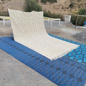 May include: A large, white, woven rug with a textured pattern is being laid out on two blue patterned rugs. The rugs are on a gravel surface.