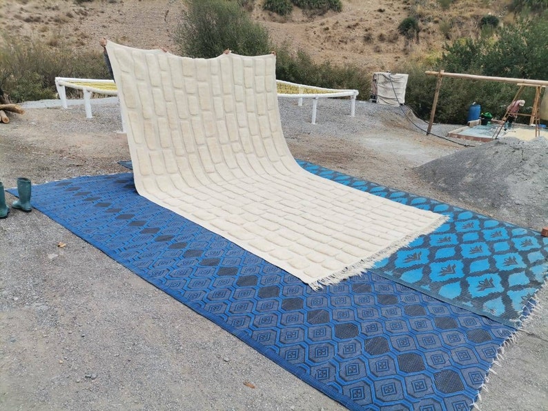 May include: A large, white, woven rug with a textured pattern is being laid out on two blue patterned rugs. The rugs are on a gravel surface.