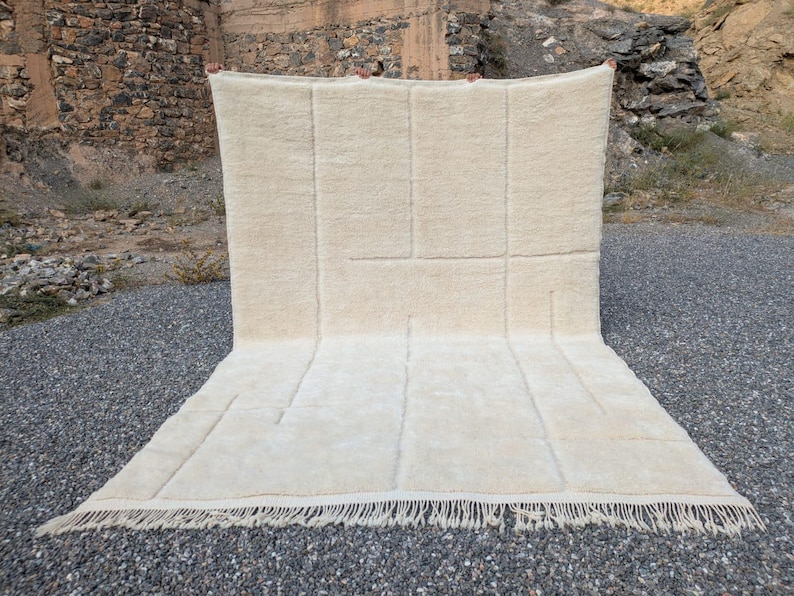 May include: A large, rectangular cream-colored rug with a geometric design. The rug features a series of rectangular and linear patterns, with a fringe along the bottom edge. The rug is made of a soft, textured material.