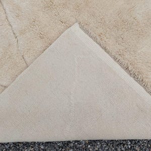 May include: Beige area rug with a plush, textured surface. The underside of the rug is a lighter beige with a woven pattern. The rug's corner is folded back, revealing the backing. The rug is on a dark surface.
