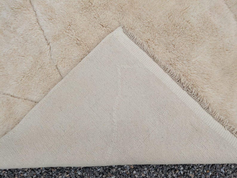 May include: Beige area rug with a plush, textured surface. The underside of the rug is a lighter beige with a woven pattern. The rug's corner is folded back, revealing the backing. The rug is on a dark surface.