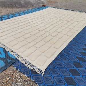 May include: A white, rectangular rug with a textured brick pattern. The rug has a fringe border and is lying on a blue and black patterned rug.
