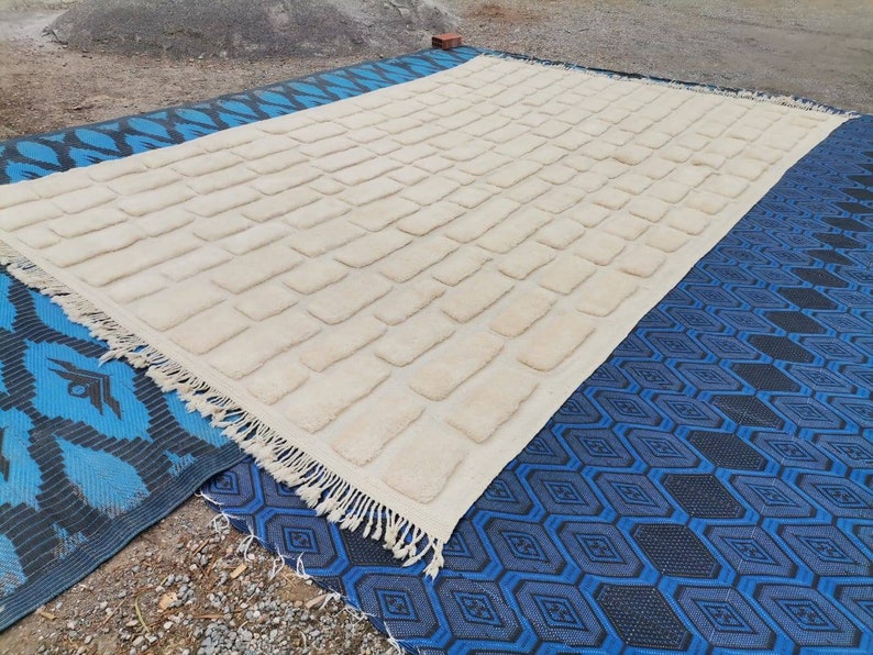 May include: A white, rectangular rug with a textured brick pattern. The rug has a fringe border and is lying on a blue and black patterned rug.