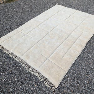 May include: Cream-colored rectangular rug with a geometric design. The rug has a textured surface with raised rectangular and linear patterns. The edges are finished with a fringe. The rug is placed on a dark gray surface.