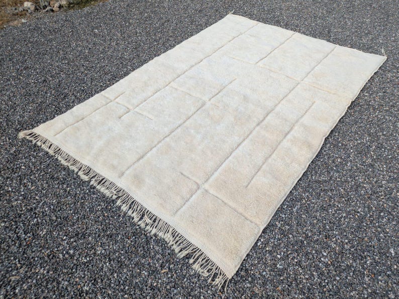 May include: Cream-colored rectangular rug with a geometric design. The rug has a textured surface with raised rectangular and linear patterns. The edges are finished with a fringe. The rug is placed on a dark gray surface.