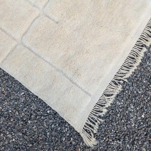 May include: Cream-colored, rectangular rug with a geometric design. The rug has a soft, textured surface and a fringe detail along one edge. The rug is partially rolled up, revealing the underside.