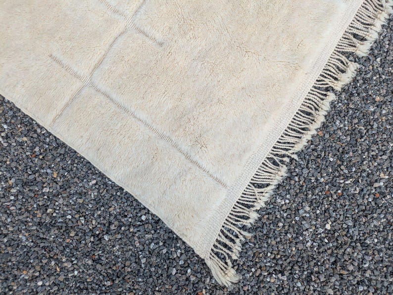 May include: Cream-colored, rectangular rug with a geometric design. The rug has a soft, textured surface and a fringe detail along one edge. The rug is partially rolled up, revealing the underside.