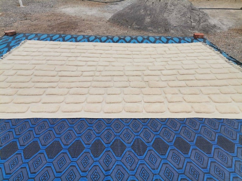 May include: A cream-colored rug with a textured, brick-like pattern. The rug is laid out on a blue and black patterned surface.