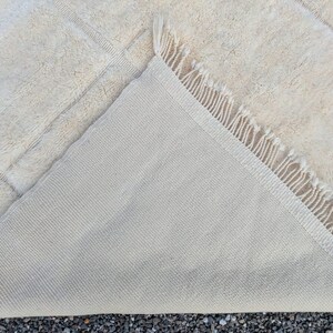 May include: Cream-colored rectangular rug with a geometric pattern. The rug's underside is visible, revealing a lighter, woven backing. The edge features a fringe detail. The rug is made of a soft, textured material.