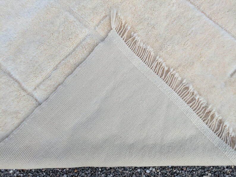 May include: Cream-colored rectangular rug with a geometric pattern. The rug's underside is visible, revealing a lighter, woven backing. The edge features a fringe detail. The rug is made of a soft, textured material.