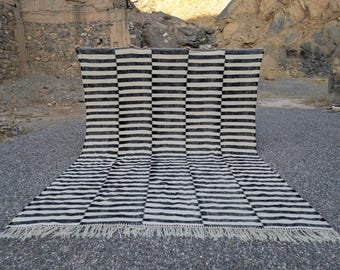 Puede incluir: Una alfombra a rayas blancas y negras con un patrón geométrico. La alfombra tiene flecos a lo largo del borde inferior. La alfombra se exhibe al aire libre sobre una superficie de grava, con una pared de piedra y terreno rocoso al fondo.