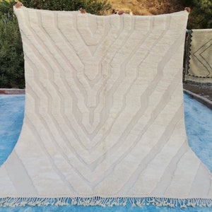 May include: A white woven rug with a geometric pattern. The rug has a fringe border and is hanging on a clothesline.