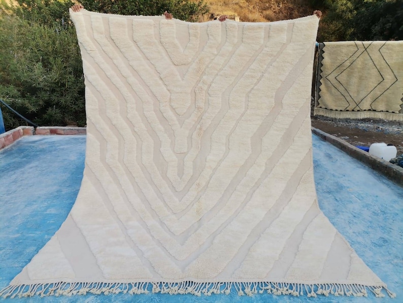 May include: A white woven rug with a geometric pattern. The rug has a fringe border and is hanging on a clothesline.