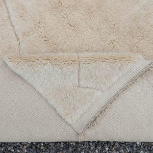 May include: A beige rug with a thick, shaggy pile. The rug's underside is visible, revealing a lighter, woven texture. The corner is folded over, showcasing the contrast between the textures. The rug appears to be made of natural fibers.