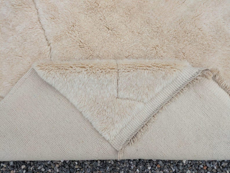May include: A beige rug with a thick, shaggy pile. The rug's underside is visible, revealing a lighter, woven texture. The corner is folded over, showcasing the contrast between the textures. The rug appears to be made of natural fibers.