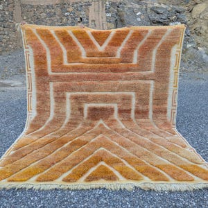 May include: A rectangular rug with a geometric pattern in shades of orange, tan, and cream. The rug features a central design with a series of lines and shapes, and a fringed edge. The rug is displayed outdoors.