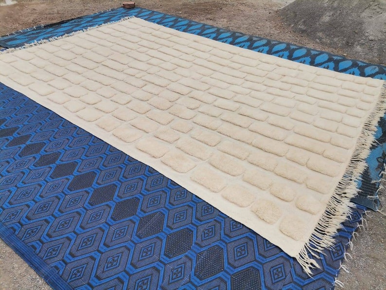 May include: A white rug with a textured brick pattern. The rug has a fringe border and is lying on a blue and black patterned rug.