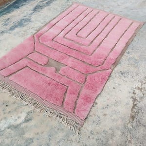 May include: A rectangular pink rug with a geometric design. The rug features a series of raised, rectangular shapes in a lighter shade of pink, creating a textured pattern. The rug has a fringe along one edge.