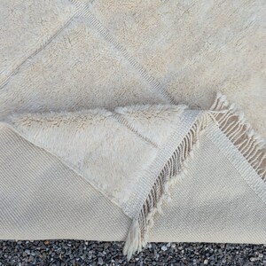 May include: Cream-colored, textured rug with a geometric diamond pattern. The rug has a thick, plush pile and a fringed edge. The underside of the rug is visible, showing a lighter, woven backing. A cozy and stylish floor covering.