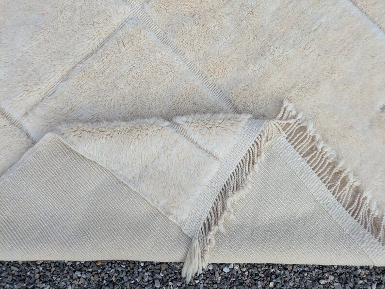 May include: Cream-colored, textured rug with a geometric diamond pattern. The rug has a thick, plush pile and a fringed edge. The underside of the rug is visible, showing a lighter, woven backing. A cozy and stylish floor covering.