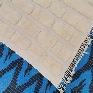 May include: A white rug with a textured brick pattern and a fringe border. The rug is laid on a blue and black patterned surface.